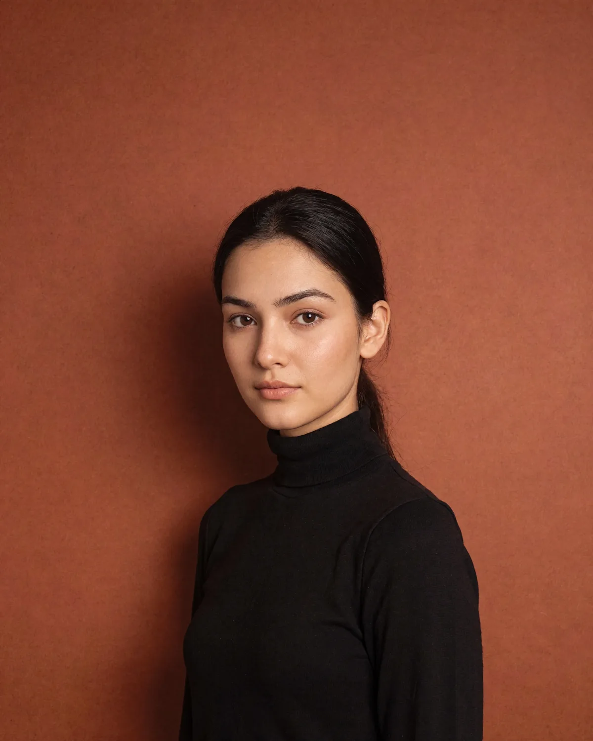 A woman in a studio against a burnt-ochre backdrop, in a black turtleneck, looking straight into the camera.
